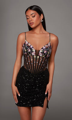 A woman wearing the Alyce 40238 Short Homecoming Dress—a fitted black sequin dress featuring a sheer floral embroidered corset bodice, thin straps, and a thigh-high slit—stands against a plain gray background, showcasing a modern twist on the classic homecoming look.