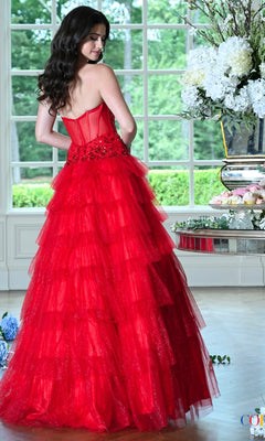 A woman wears a Colors Dress 4024 strapless red glitter tulle ball gown with a corset bodice and a tiered skirt while standing near a window.