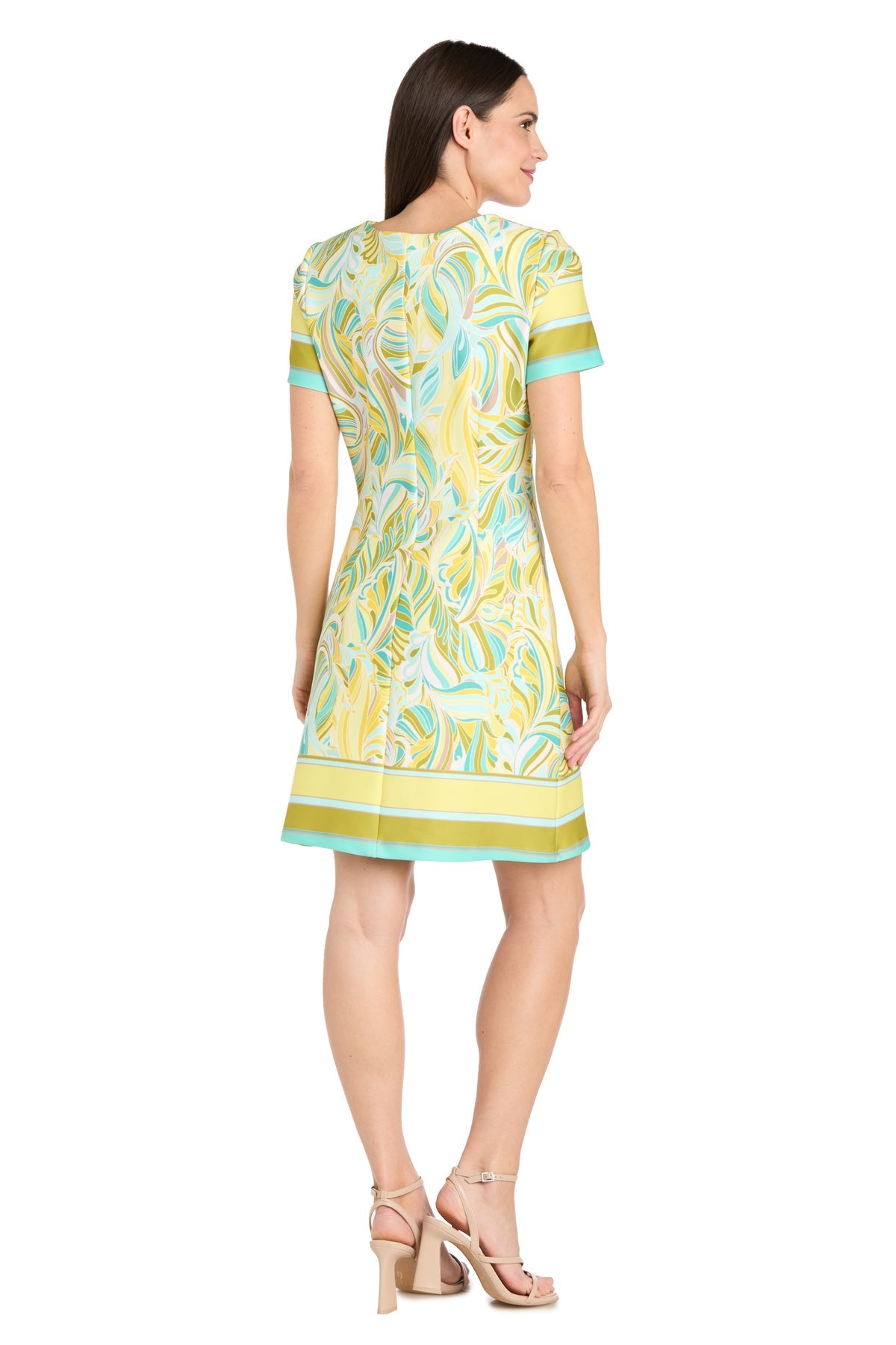 A woman stands facing away, wearing the R&M Richards 4026 short yellow print party dress with abstract green and blue patterns, paired with beige heeled sandals.