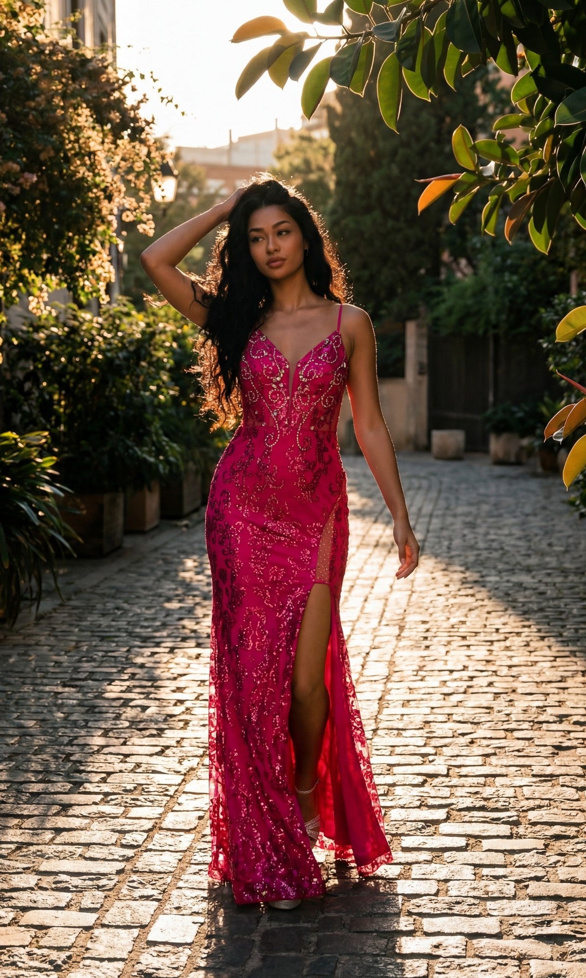 A woman wearing the Long Prom Dress: Chicas 4028, featuring a bright pink, sparkly design with a slit and sheer corset bodice, walks along a sunlit cobblestone path surrounded by greenery.
