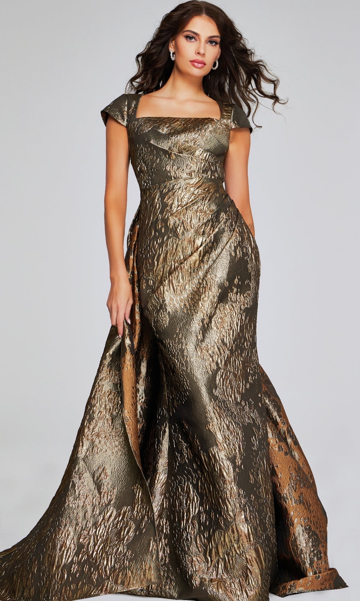 A woman wears the Jovani 40295 long formal dress, a metallic bronze and gold evening gown with cap sleeves and a square neckline, standing against a plain background.