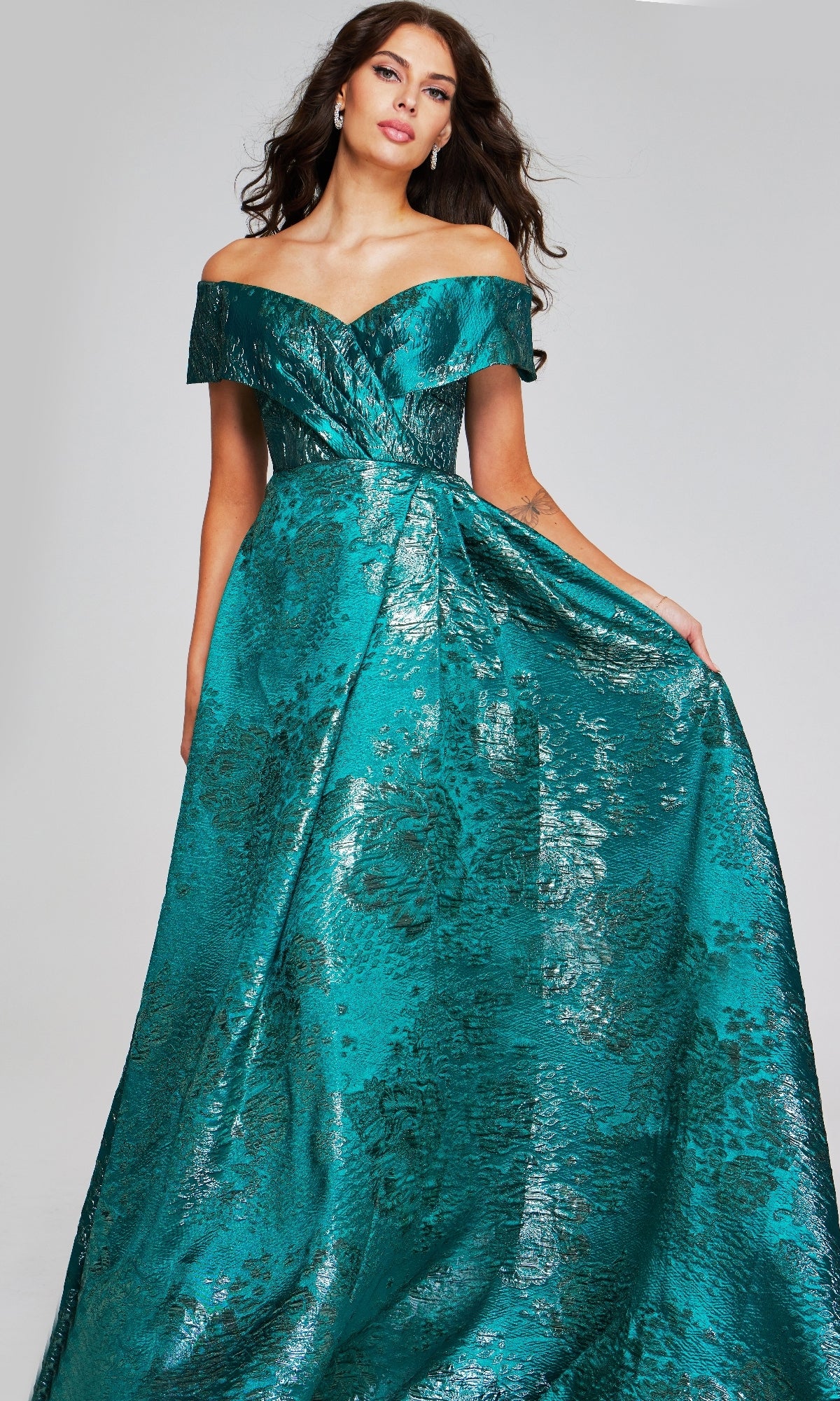 Wearing the Long Formal Dress: Jovani 40296, a woman stands against a plain background, elegantly holding part of the off-the-shoulder metallic textured gown with a floral pattern to the side.
