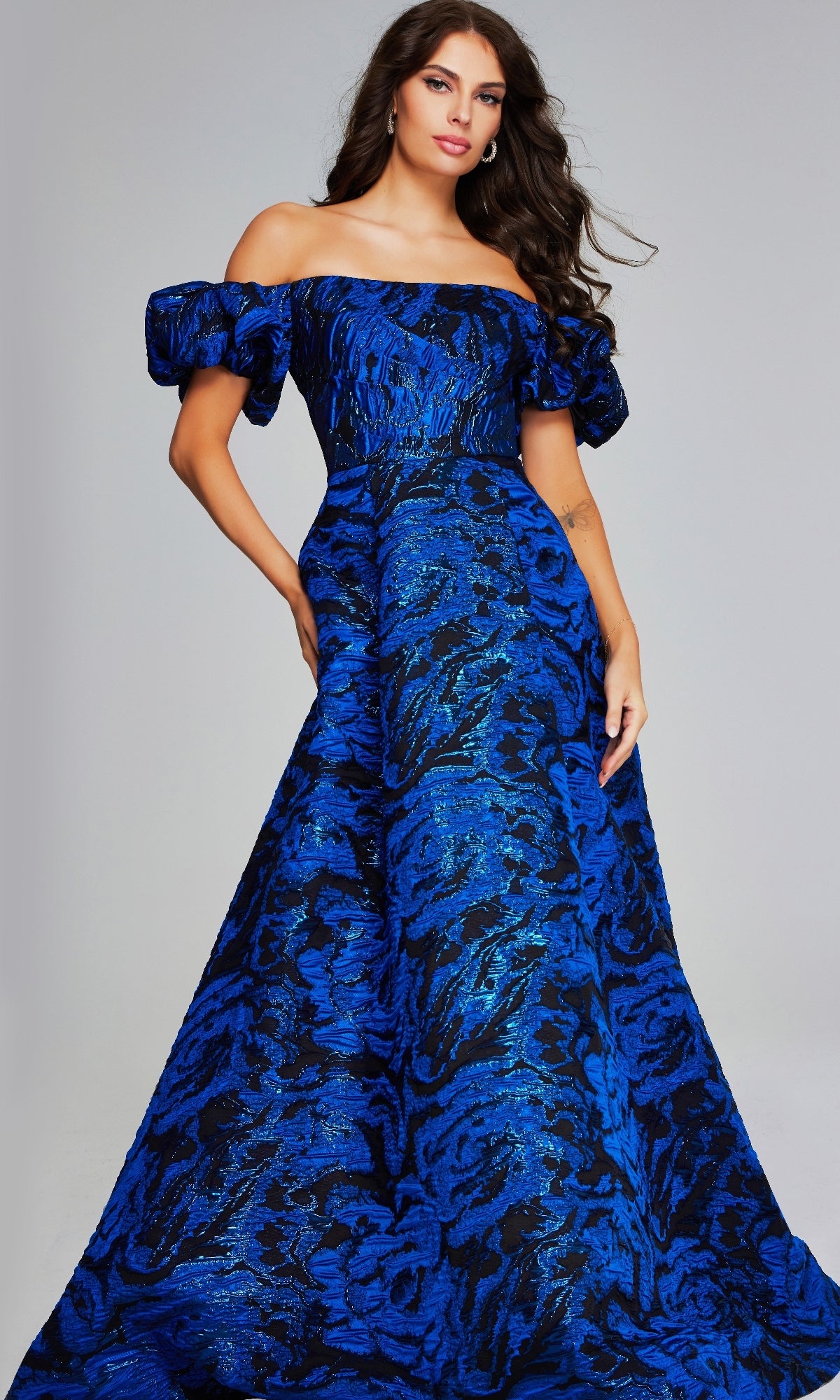 A woman stands elegantly against a plain light background, wearing the Jovani 40315 long formal dress—an off-the-shoulder ball gown with a blue and black patterned design and puff sleeves.