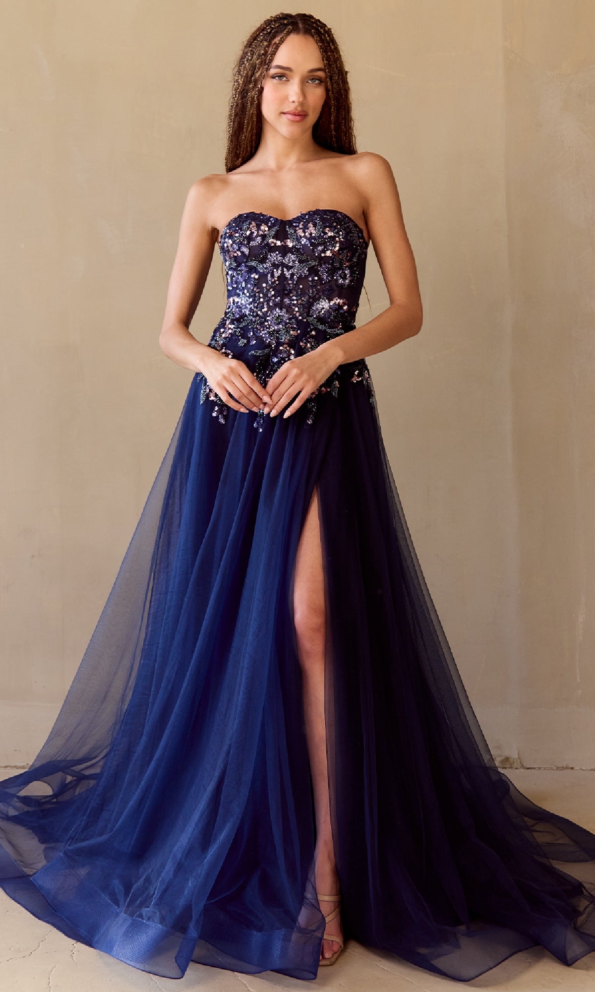 A woman poses against a plain beige wall wearing the Amelia Couture 4031 Strapless A-Line Prom Gown, featuring a sequin corset bodice, sheer tulle skirt, and high leg slit.