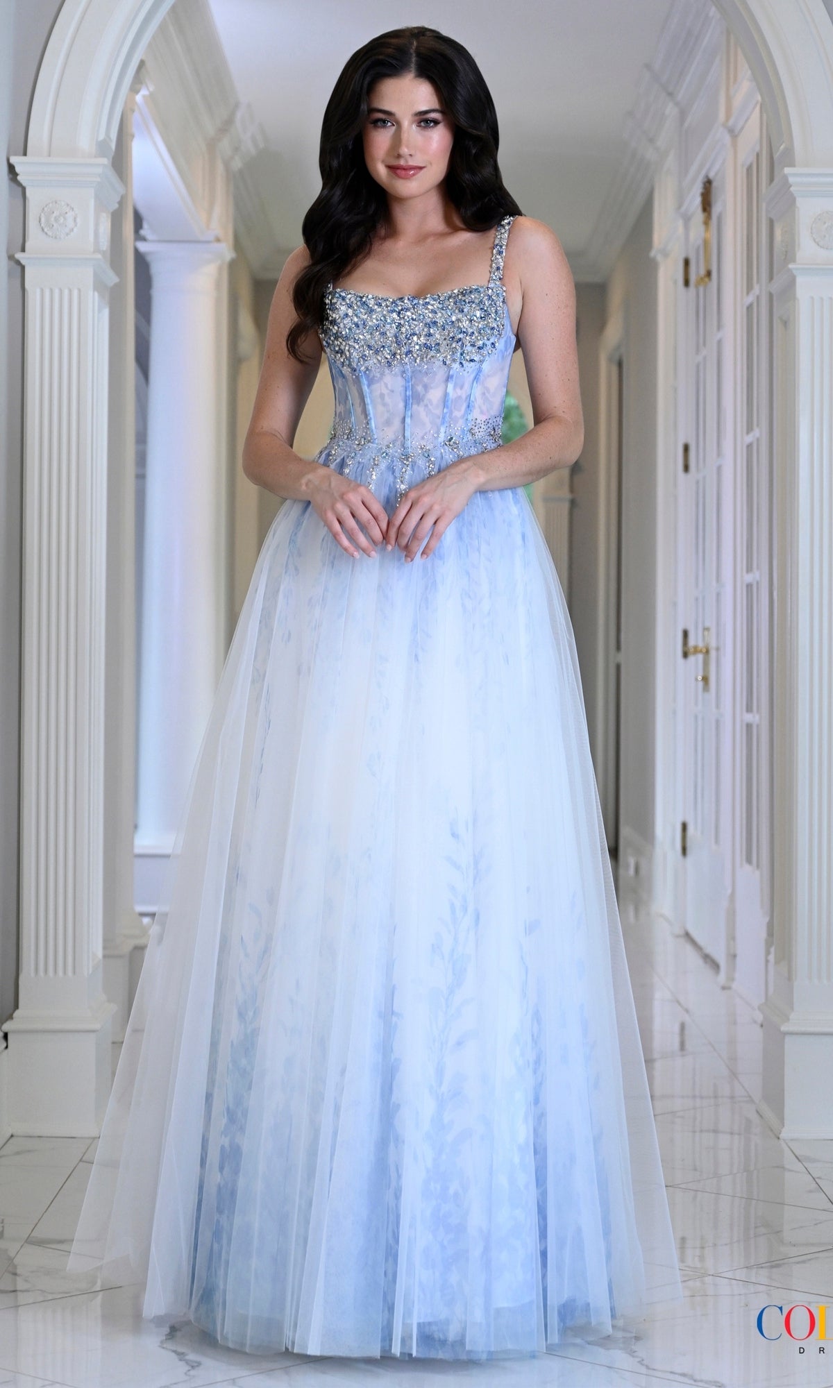 Colors Dress 4033 is a floor-length baby blue prom dress featuring a crystal beaded bodice and a sheer, patterned tulle overlay skirt.
