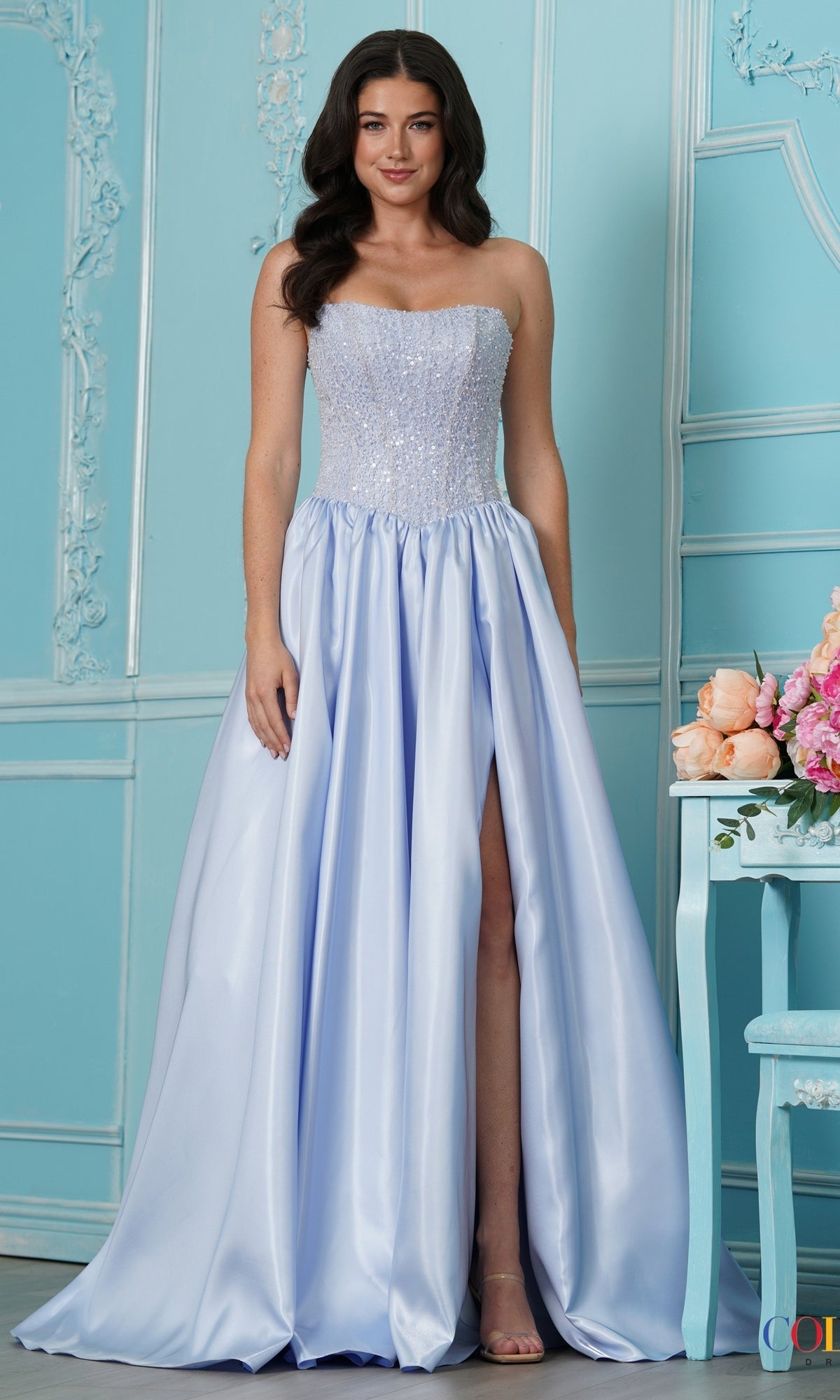 A woman wears a light blue Colors Dress 4034 strapless ball gown with a beaded bodice and a floor-length skirt featuring a high side slit.