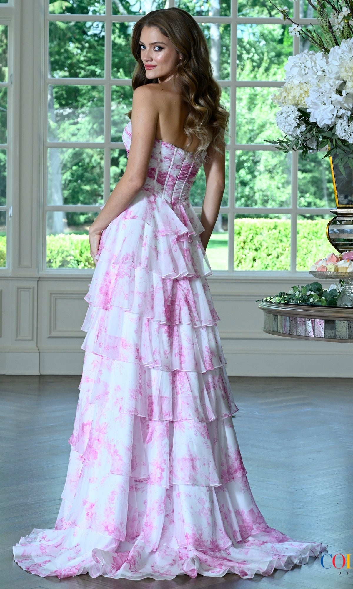 The Colors Dress 4038 is a floral prom dress with a strapless sweetheart bodice, a floor-length silhouette, and tiered ruffles in a white and pink pattern.