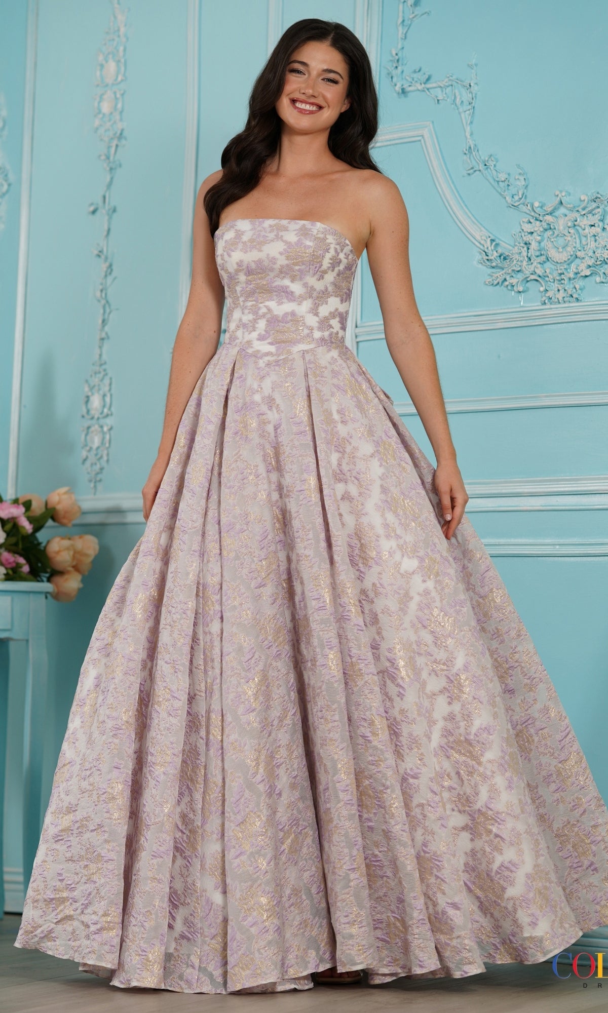 A smiling woman wears a lilac Colors Dress 4039, a strapless floral jacquard prom dress with a floor-length, full pleated skirt.