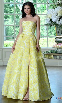 A woman wears a yellow and white floral jacquard Long Prom Dress: Colors Dress 4039, a strapless floor-length gown featuring a front slit.