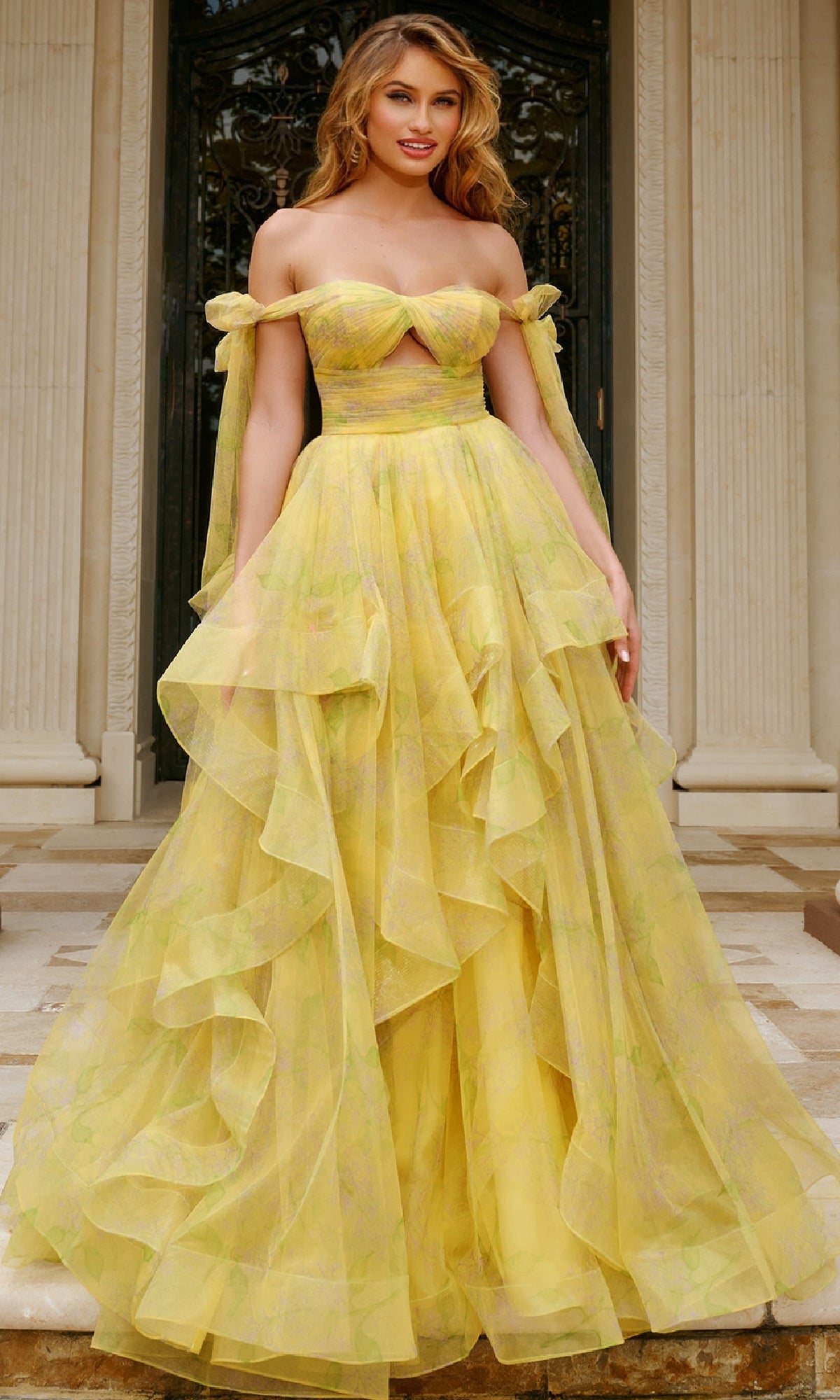 A woman stands on stone steps in front of ornate doors and columns, wearing the Long Prom Dress: Jovani 40400—a yellow off-the-shoulder gown with a layered ball-gown skirt and sheer detailing.
