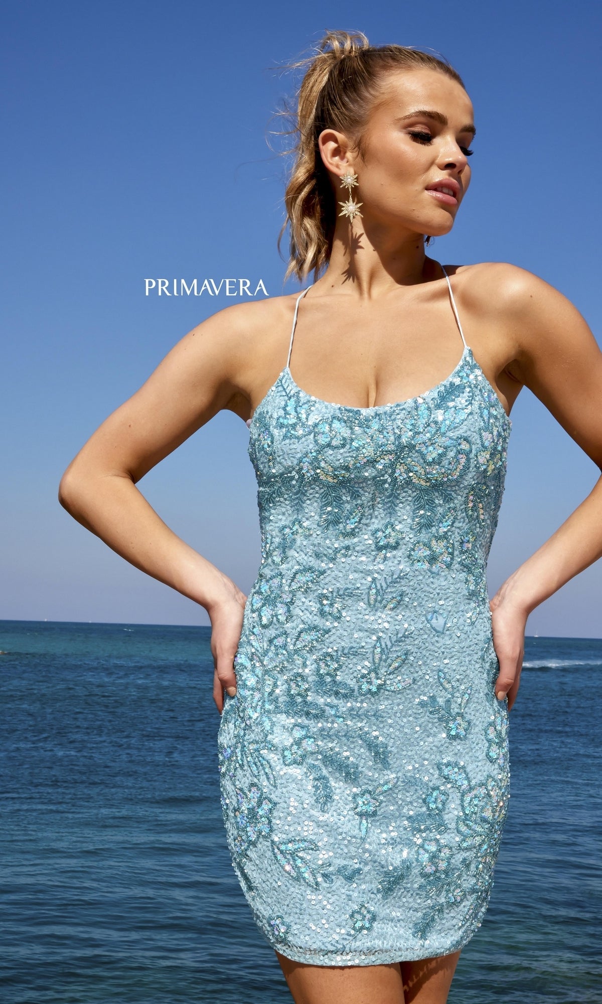 A woman stands by the ocean wearing the Lace-Up Back Short Beaded Homecoming Dress 4040 in light blue with spaghetti straps and star-shaped earrings. 