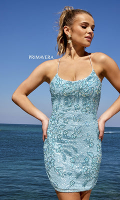 A woman stands by the ocean wearing the Lace-Up Back Short Beaded Homecoming Dress 4040 in light blue with spaghetti straps and star-shaped earrings. "PRIMAVERA" is displayed in the background.