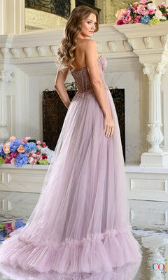 A woman wearing a Long Prom Dress: Colors Dress 4041, featuring a floor-length lavender design with a sheer lace corset bodice and a ruffled tulle skirt, standing before a floral-decorated fireplace.
