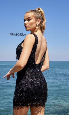 A woman in a black Beaded-Fringe Short Semi-Formal Dress 4042 stands by the ocean with her back to the camera, hair in a high ponytail, under a clear blue sky. The word "PRIMAVERA" appears in the scene.