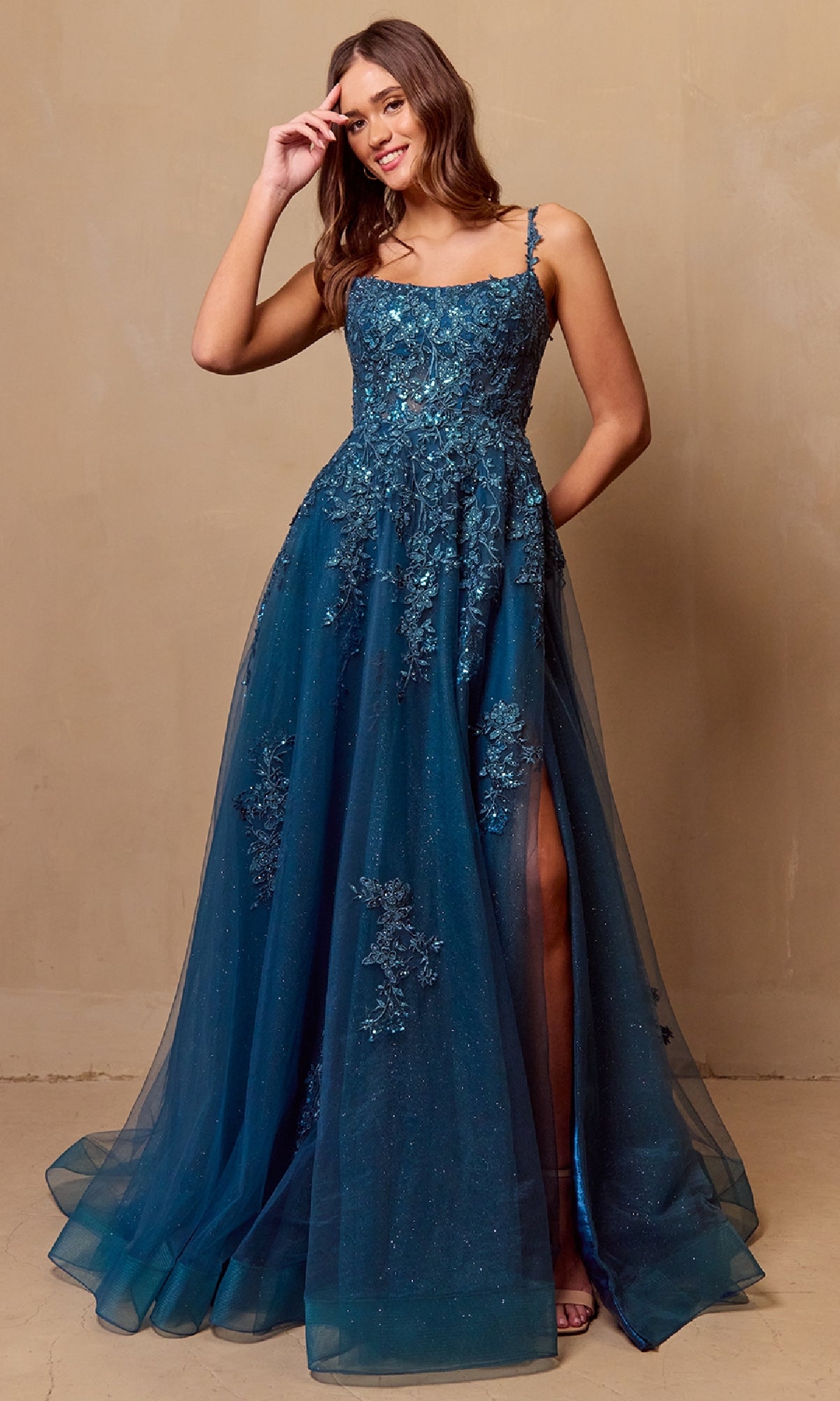 A woman stands against a plain beige background wearing the Amelia Couture 4044, a blue, floor-length corset prom ball gown featuring an A-line silhouette, sleeveless design, floral embellishments, and a side slit.