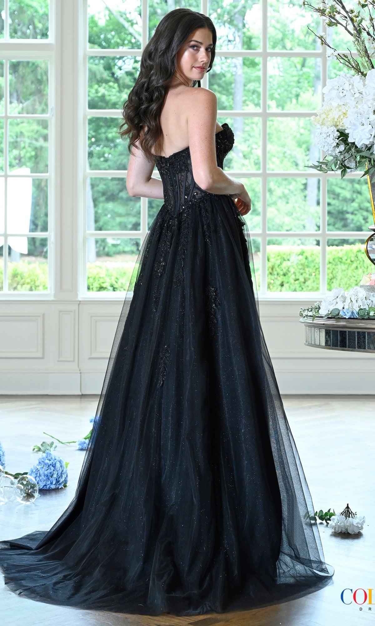 A person wears a Long Prom Dress: Colors Dress 4044 featuring a black corset, strapless bodice, and a sheer, embellished tulle skirt.
