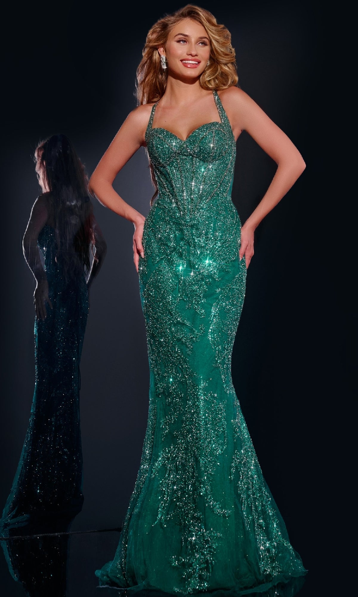 A woman poses in the Jovani 40470 Lace-Up Long Beaded Prom Dress in emerald, featuring a fitted silhouette and straps, against a dark backdrop with her reflection visible.