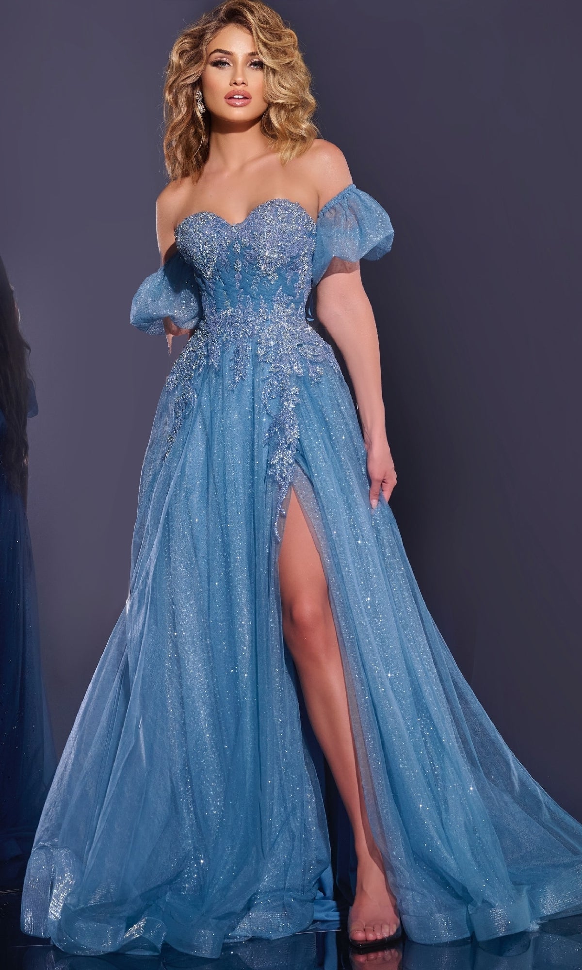 A woman poses against a dark background in the Long Blue Prom Dress: Jovani 40474, featuring a shimmering fabric, sweetheart neckline, puff sleeves, floral embellishments, and a high slit.