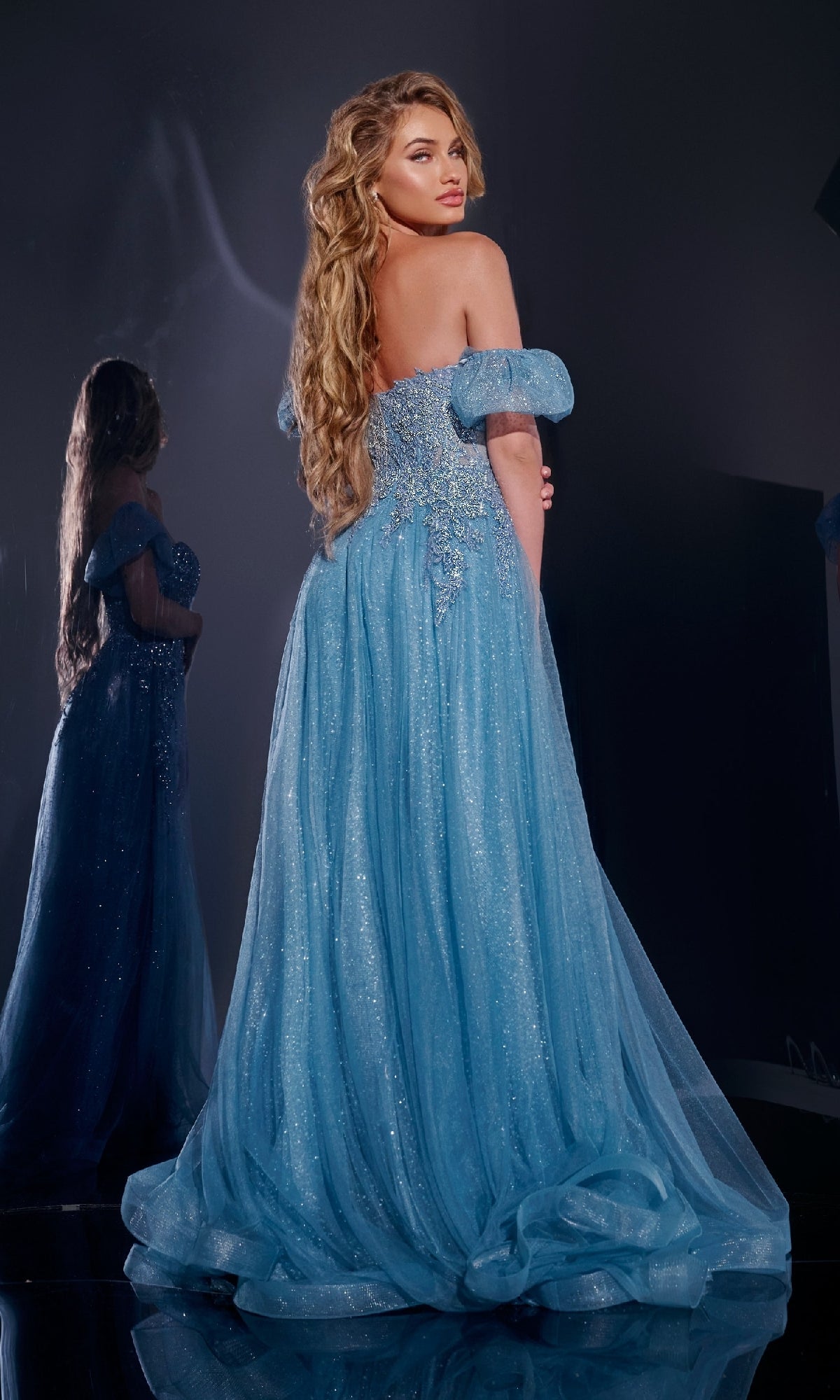 A woman with long, wavy hair models the Jovani 40474 long blue prom dress, standing before a mirror to highlight the off-the-shoulder sparkling design and stunning back of the gown.