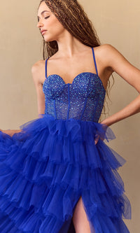 Woman wearing a royal blue dress with a sparkly corset bodice and a tiered ruffle ball gown skirt standing in front of a beige background.