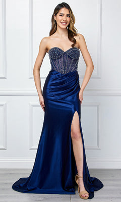 A woman in the Amelia Couture 4049 strapless long prom gown, featuring a navy blue beaded bodice and thigh-high slit, paired with nude heels, stands before a white paneled wall.