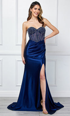 A woman stands against a white-paneled wall wearing the Strapless Long Prom Gown: Amelia Couture 4049 in navy blue satin, featuring a sweetheart neckline, beaded corset bodice, thigh-high slit, and nude high-heeled sandals.