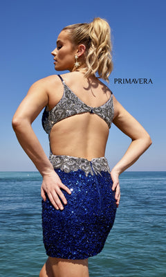 A blonde woman with a ponytail stands by the ocean wearing the Side-Beaded Short Sequin Party Dress 4052 in blue and silver with an open back. "PRIMAVERA" appears near her head.