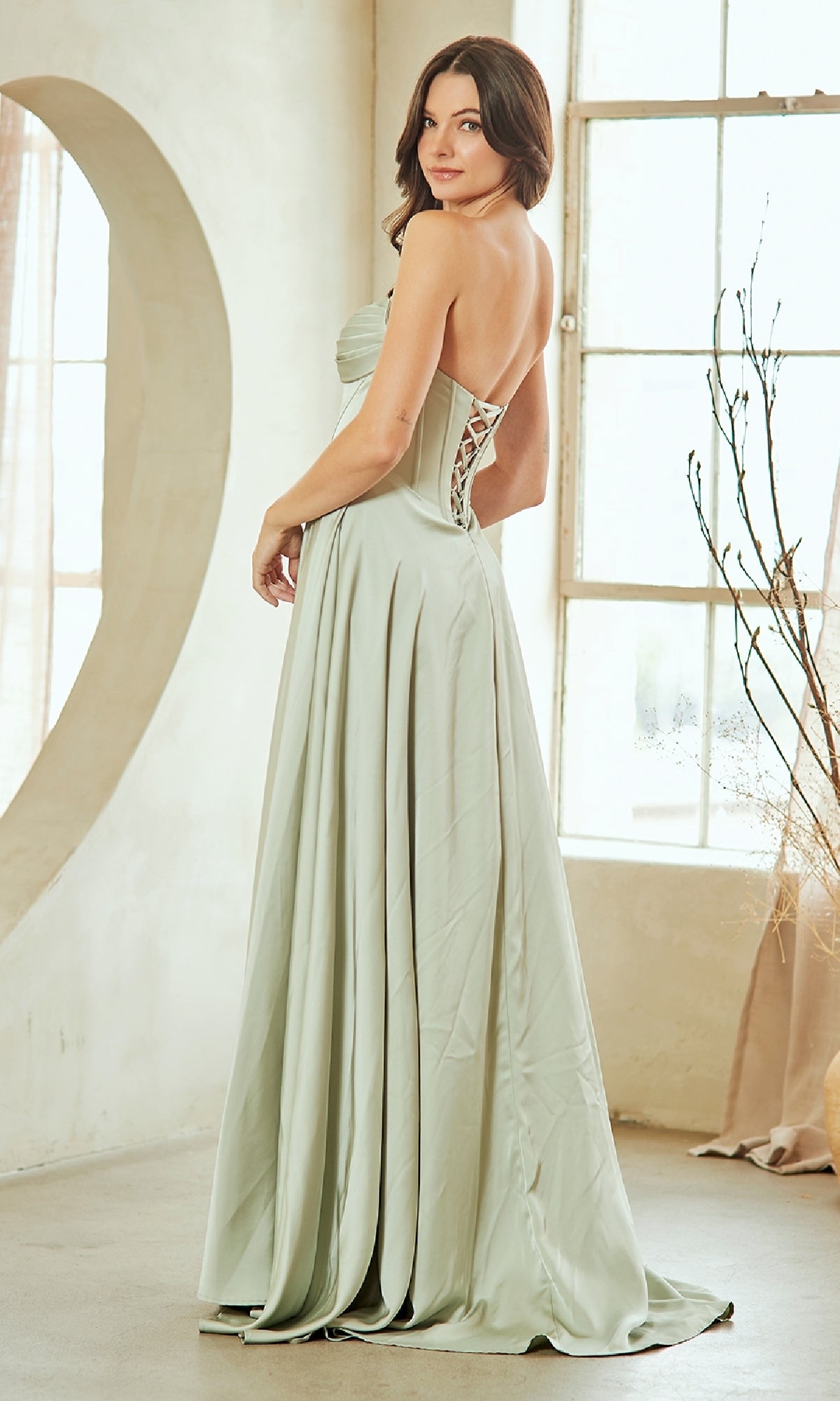 Woman showing the back of a light green evening gown with a lace-up design, standing in a bright room.