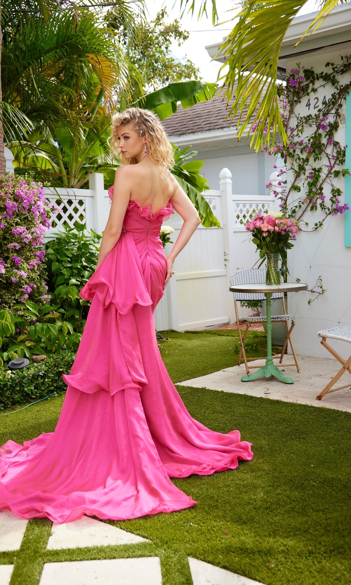 A woman stands outdoors on green grass near a white fence, wearing the Side-Sash Strapless Prom Gown: Ava Presley 40532, with flowers and a small table in the background.