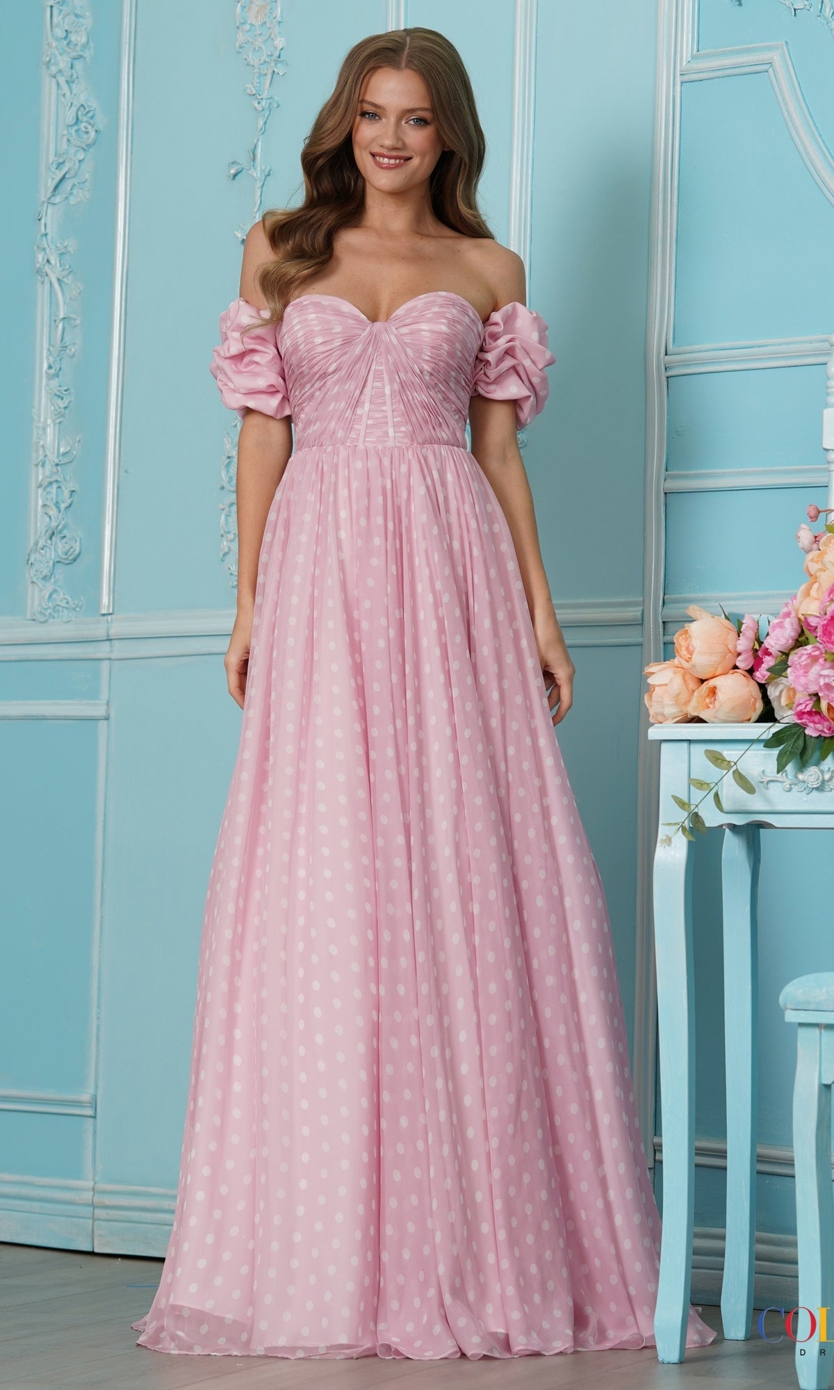 A woman wears a vintage-inspired, floor-length pink off-the-shoulder Long Prom Dress: Colors Dress 4053 with white polka dots and puffed sleeves.