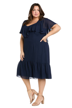 A woman models the R&M Richards 4054W Plus-Size Navy Party Dress, featuring ruffle details and a knee-length cut, styled with nude block-heeled sandals. She stands confidently with one hand on her hip.