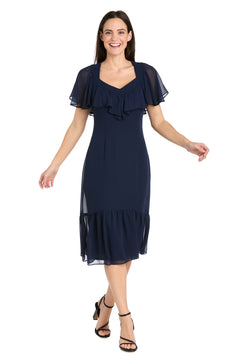 A woman stands against a white background wearing the R&M Richards 4054P Petite Navy Blue Midi Party Dress with short sleeves, ruffle details, and black strappy heels.