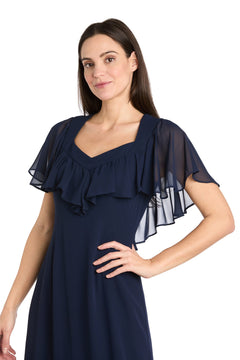 Woman modeling the R&M Richards 4054P Petite Navy Blue Midi Party Dress with sheer, ruffled sleeves, posing with one hand on her hip against a white background.