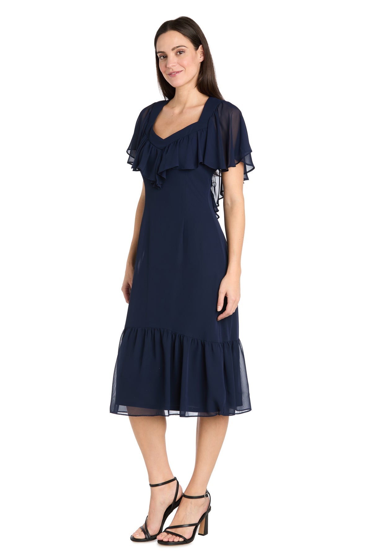 A woman models the R&M Richards 4054P Petite Navy Blue Midi Party Dress with ruffled sleeves and hem, paired with black high-heeled sandals, standing against a white background.