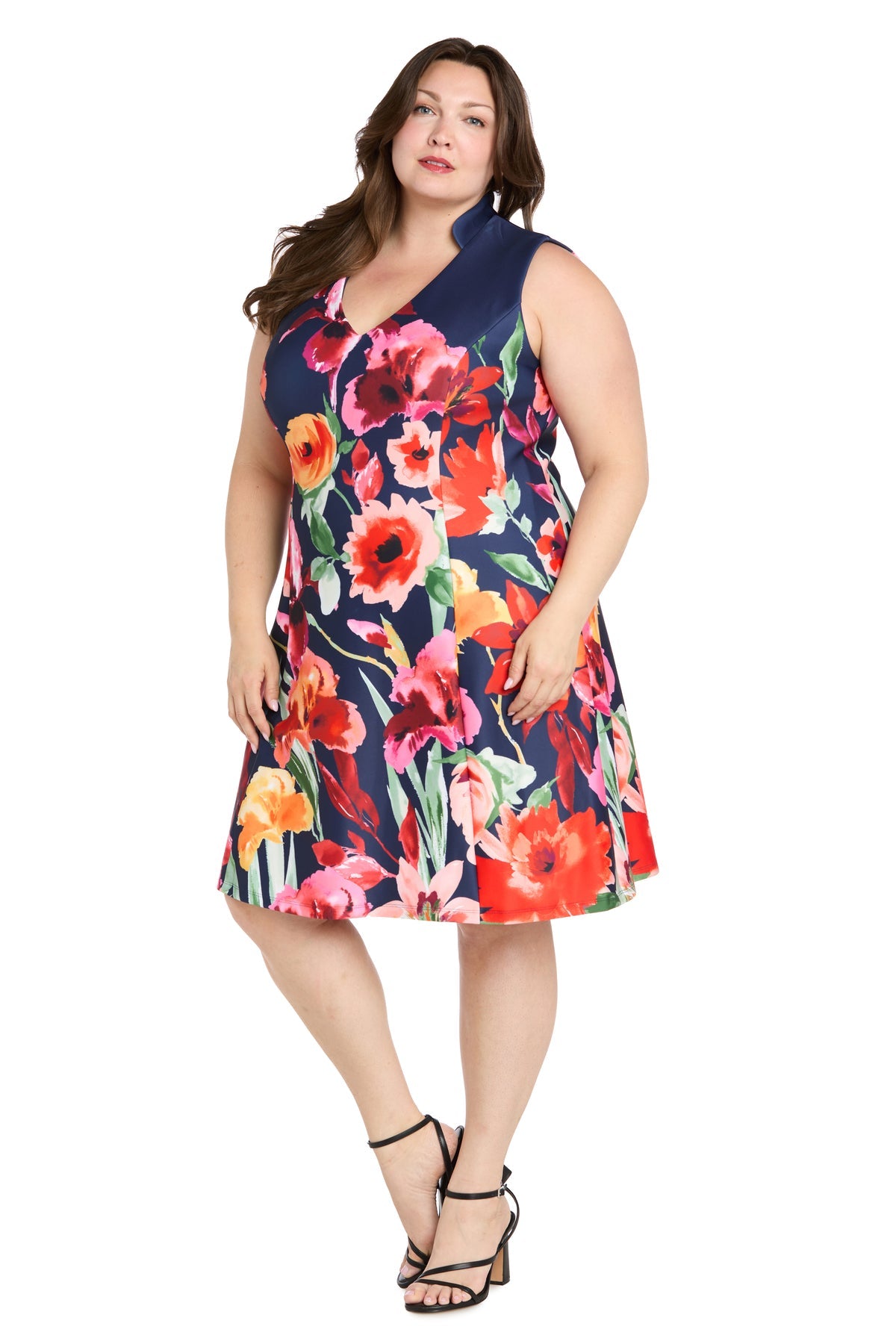 A woman models the R&M Richards 4056W Plus-Size Wedding Guest Dress, a sleeveless, knee-length floral dress, paired with black strappy heels against a white background.