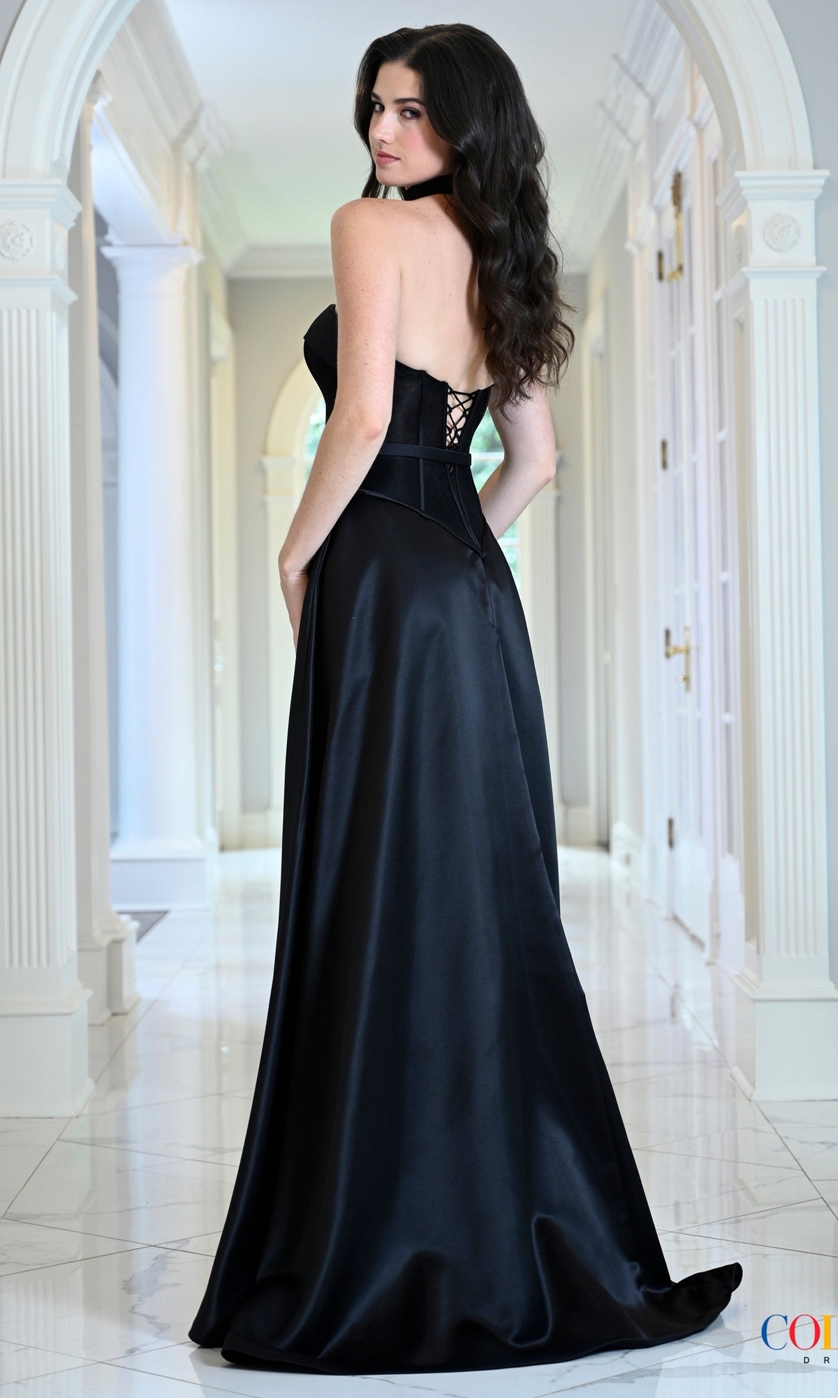 A woman wearing a Black Satin Prom Dress: Colors Dress 4056, a floor-length black corset gown with a strapless bodice and lace-up back.