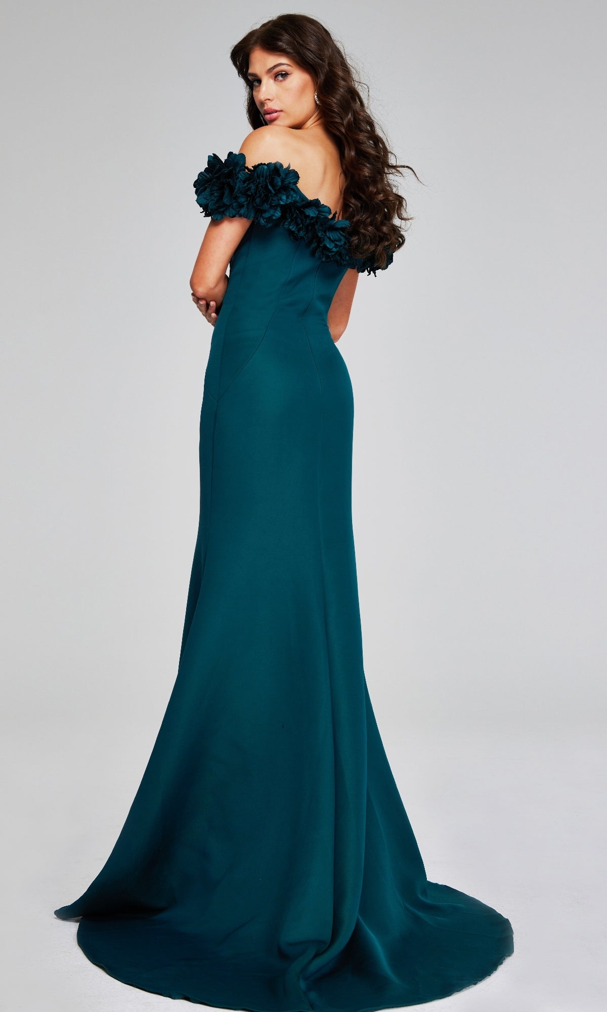A woman in the Jovani 40595 long formal dress, featuring teal off-the-shoulder style and floral-detailed sleeves, stands facing backward and looks over her shoulder.