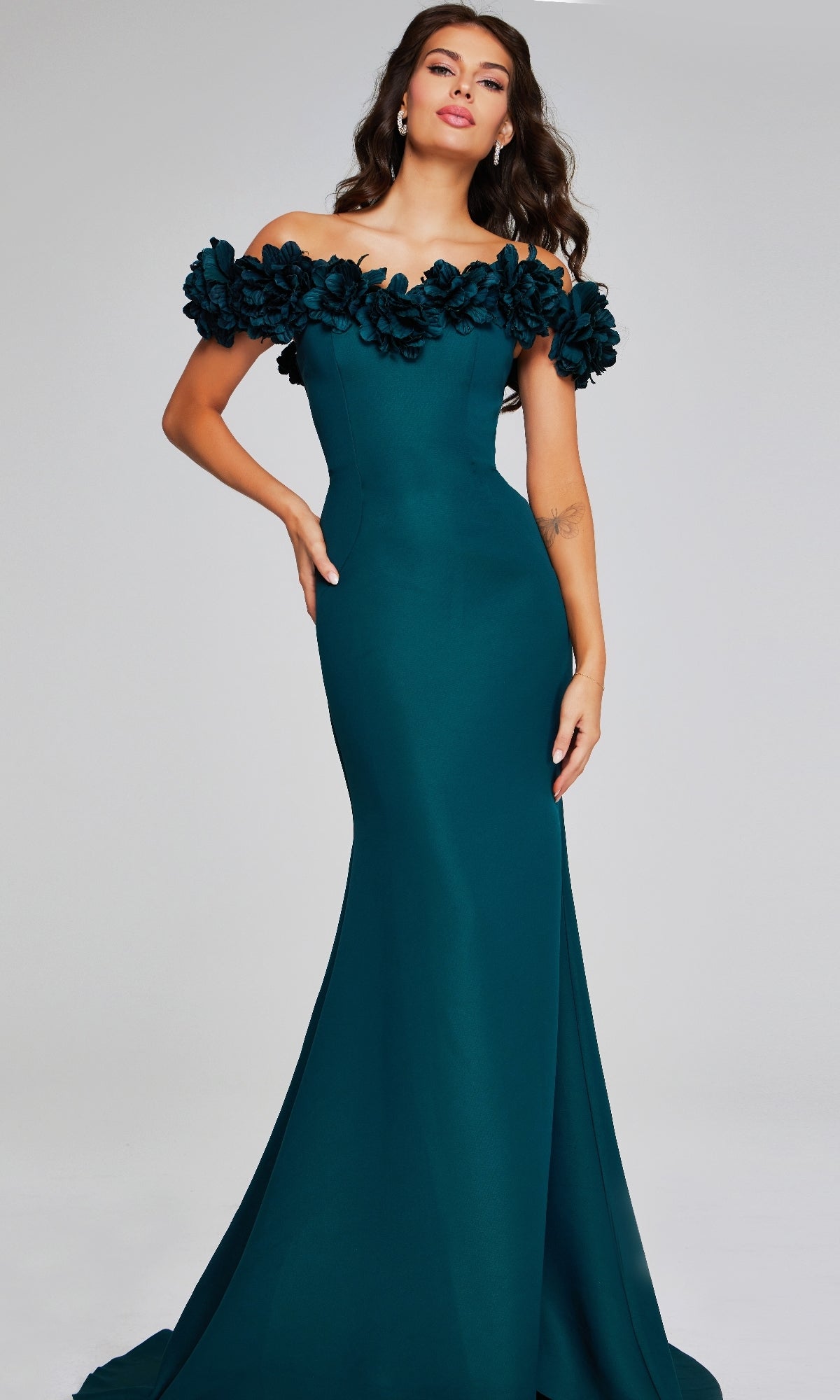 A woman stands against a plain light background wearing the Jovani 40595, a long formal teal evening dress with an off-the-shoulder neckline and floral detailing.