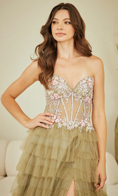 A woman wearing the Long Prom Dress: Amelia Couture 4060, an olive green strapless gown with a ruffled skirt, leg slit, and floral-embroidered semi-sheer bodice, stands indoors.