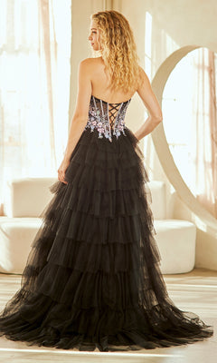 A woman with curly blonde hair stands in a sunlit room wearing the Amelia Couture 4060 Long Prom Dress—a black strapless tulle gown with floral details, a lace-up back, tiered skirt, and dramatic leg slit.