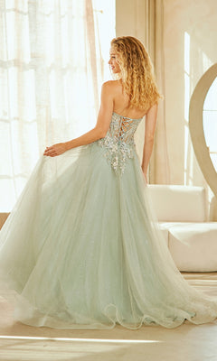 A woman in the Strapless Glitter Prom Gown: Amelia Couture 4062 with 3-D floral appliqué and lace-up back stands by a window, gracefully holding the skirt.