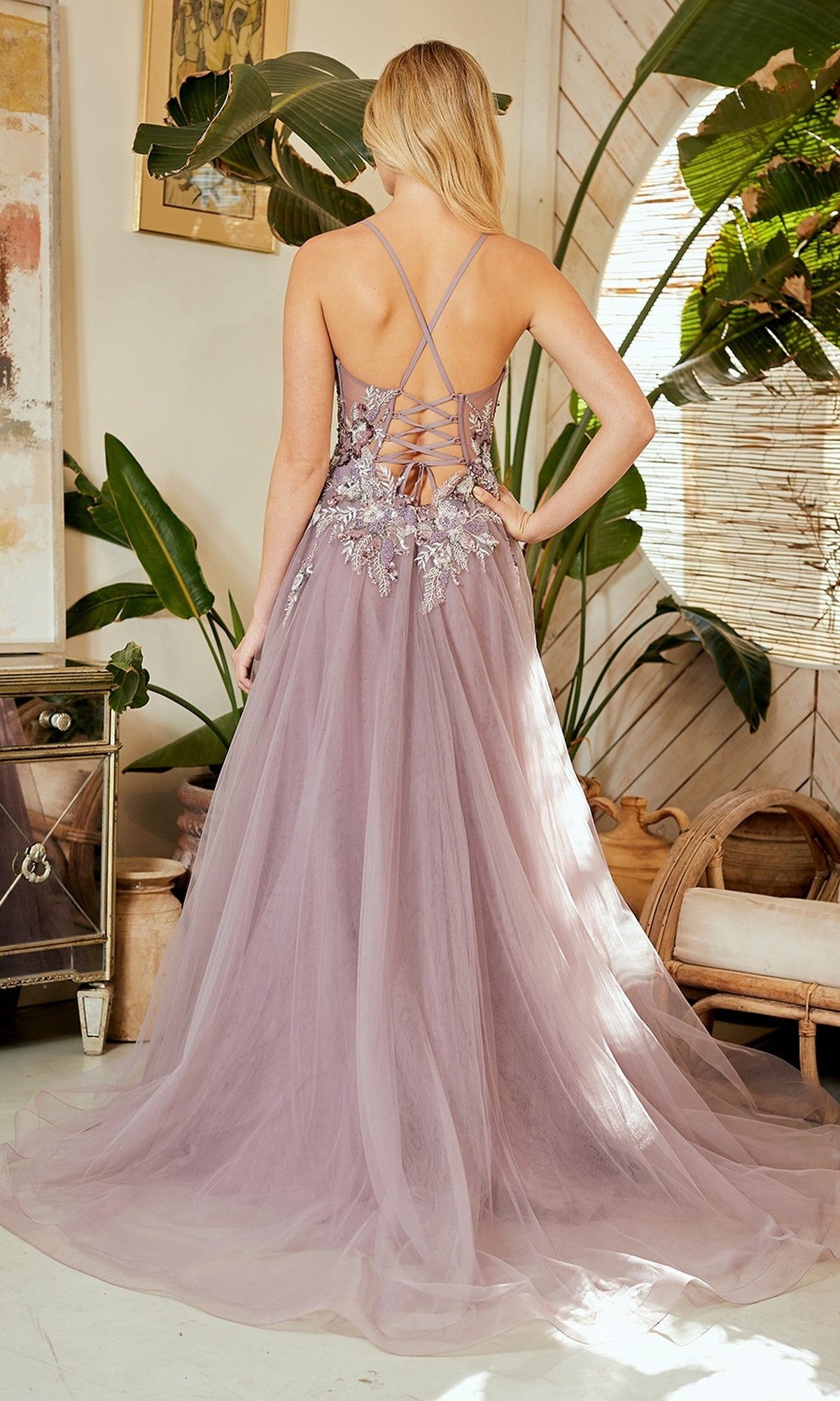 A woman stands indoors wearing the Amelia Couture 4064 long prom dress, featuring mauve tulle, a flowing A-line skirt, floral lace accents, and stylish crisscross back straps.