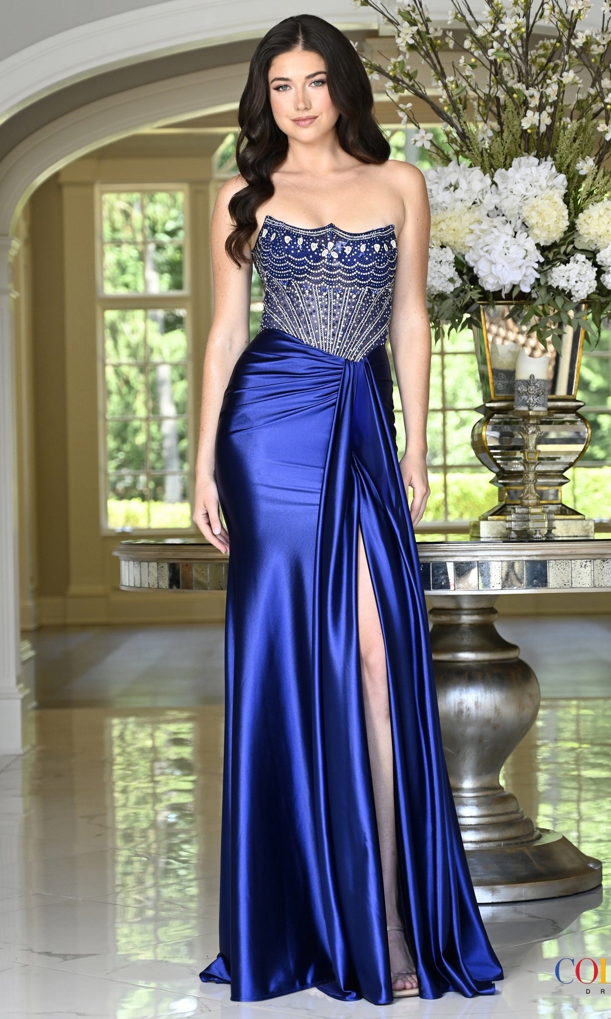 A woman wears a floor-length, deep blueberry Colors Dress 4080 corset prom dress featuring a crystal-embellished bodice and a side slit.