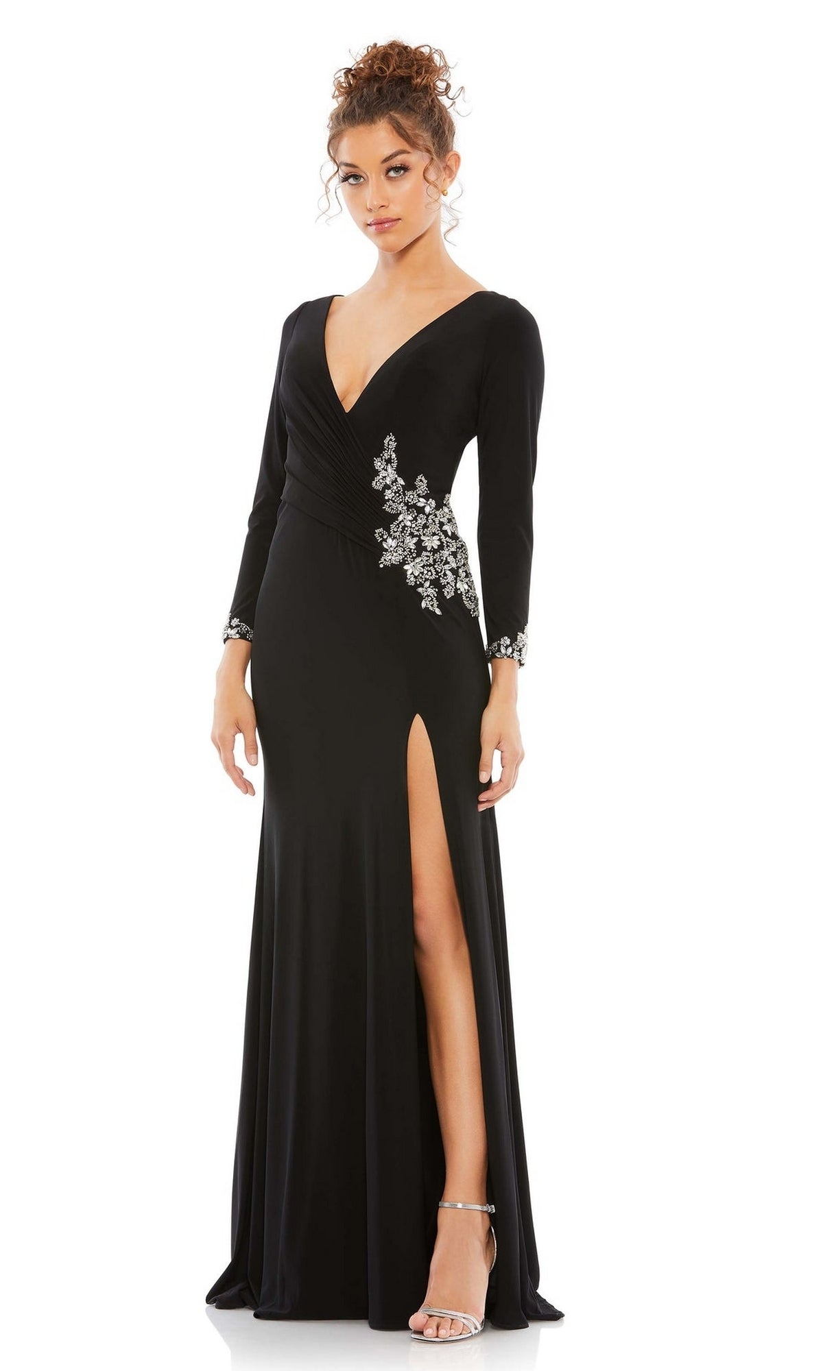 A woman wears the Long Formal Dress 41016 by Mac Duggal, featuring a fitted black design with a deep V-neck, silver floral embellishments, long sleeves, and a high leg slit, styled with silver high-heeled sandals.