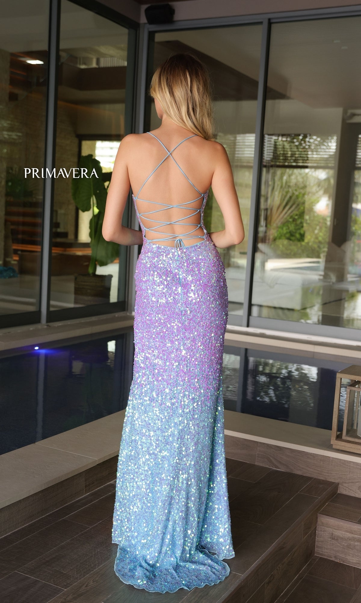 A woman stands indoors by a pool, wearing the Primavera Ombre Long Beaded Prom Dress 4102 in sparkly blue and purple with crisscross straps and an open back.