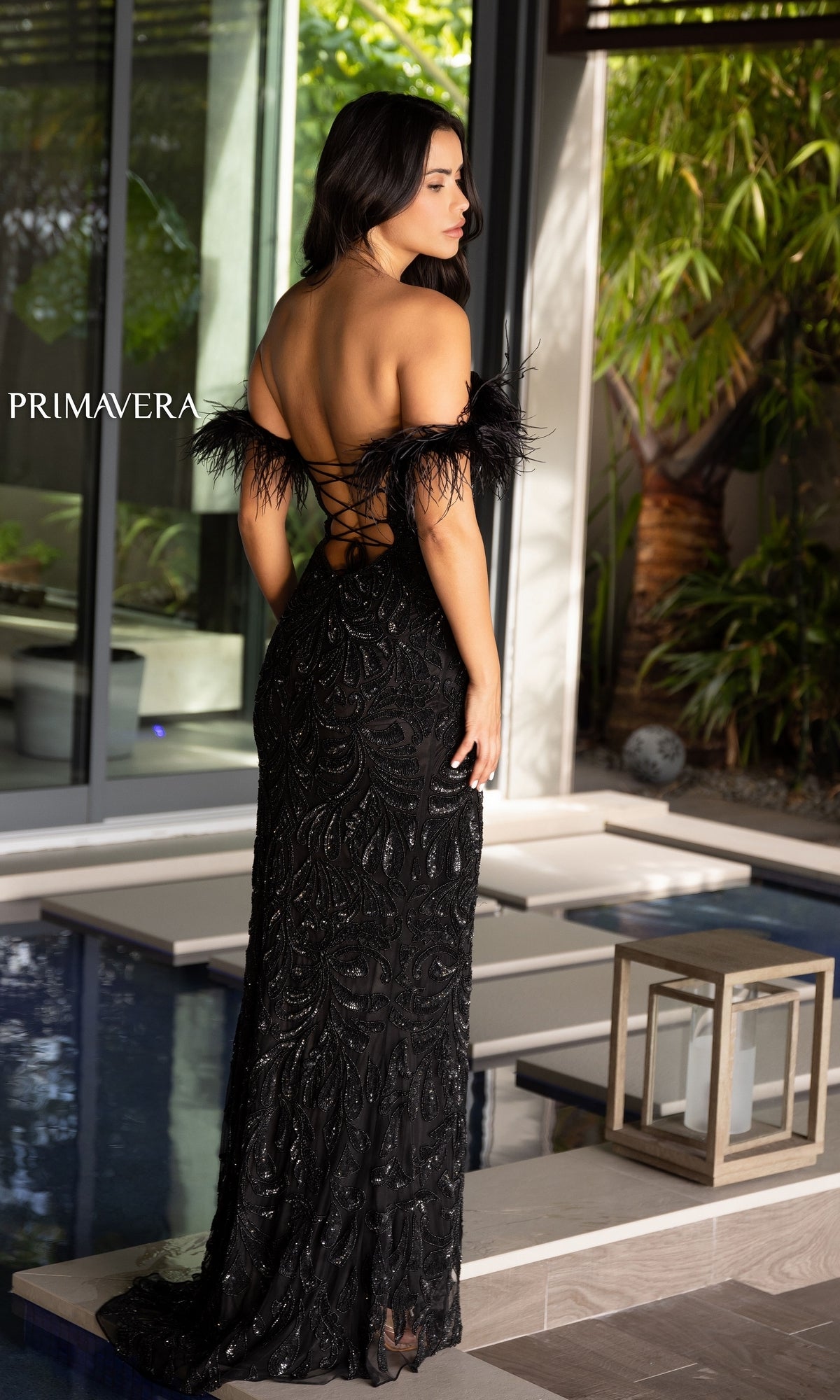 A woman in the Primavera Feather Off-Shoulder Long Prom Gown 4105, featuring feathered sleeves and sequin details, stands by a modern indoor pool facing away from the camera.