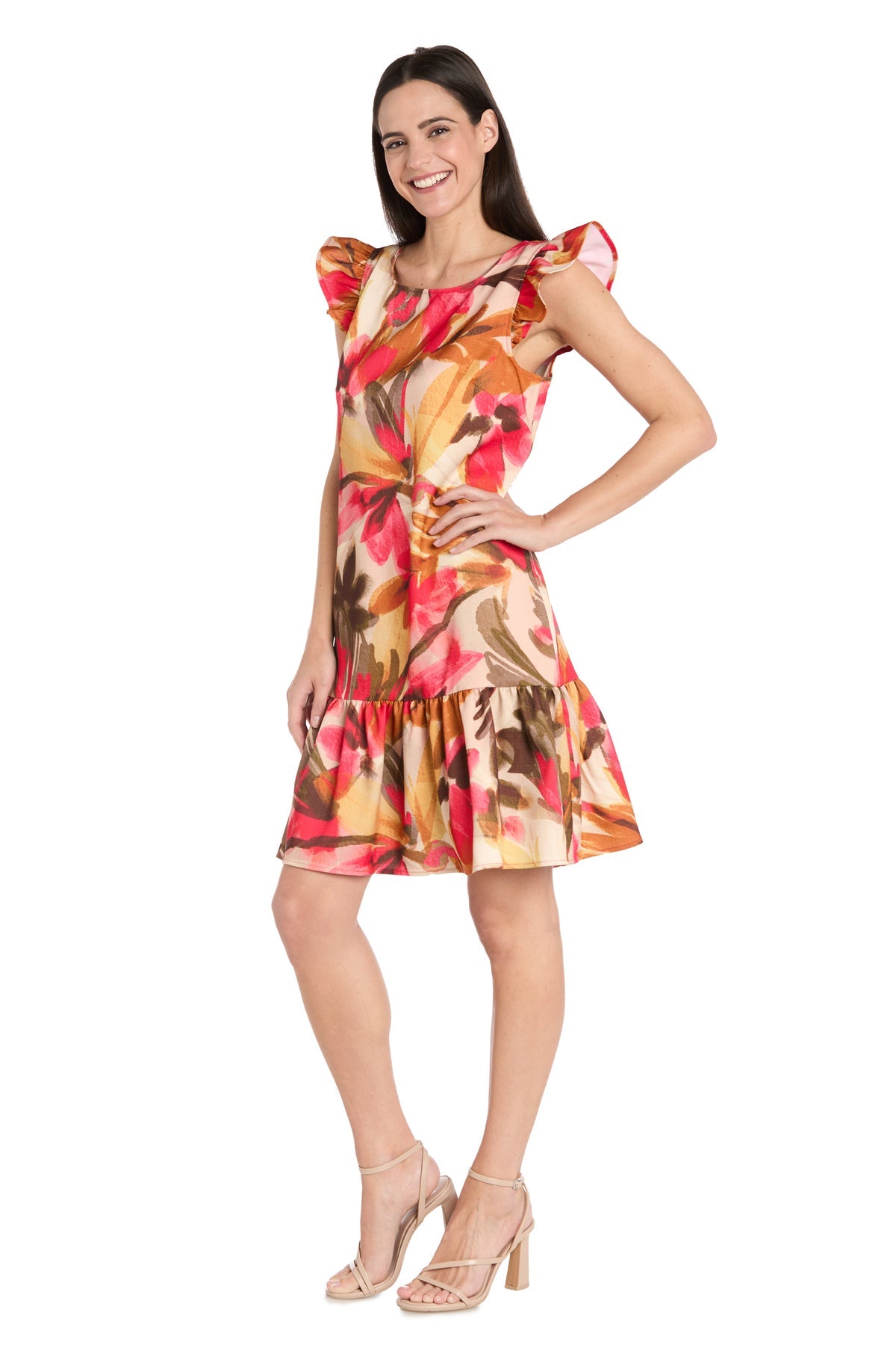 A woman stands and smiles against a white background wearing the R&M Richards 4105P Petite Short Print Party Dress with ruffled details and vibrant floral print, paired with beige high-heeled sandals.