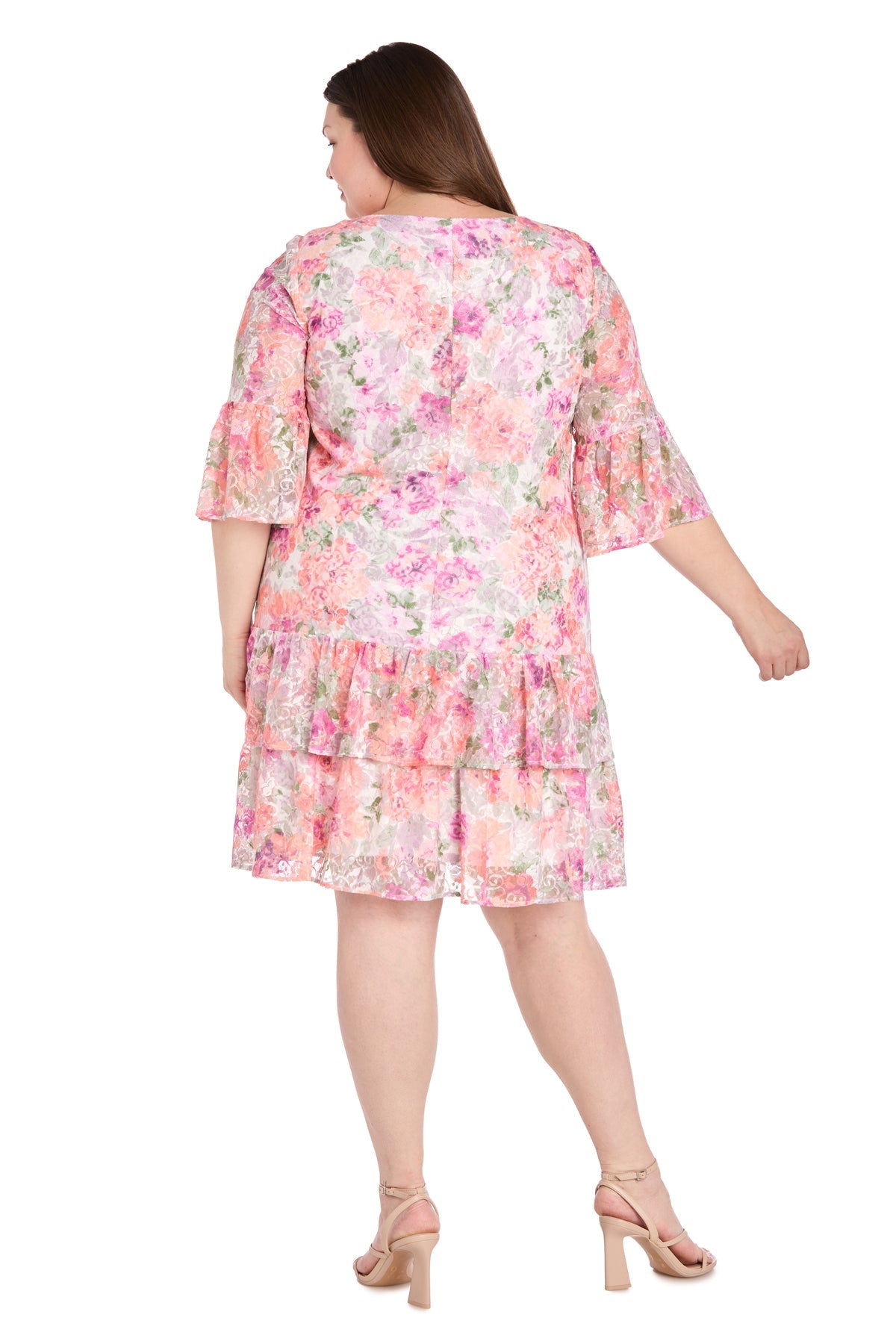 A woman models the R&M Richards 4107W Plus-Size Print Party Dress, featuring a knee-length floral design with ruffled sleeves and hem. She stands facing away to showcase the back, paired with nude heels.