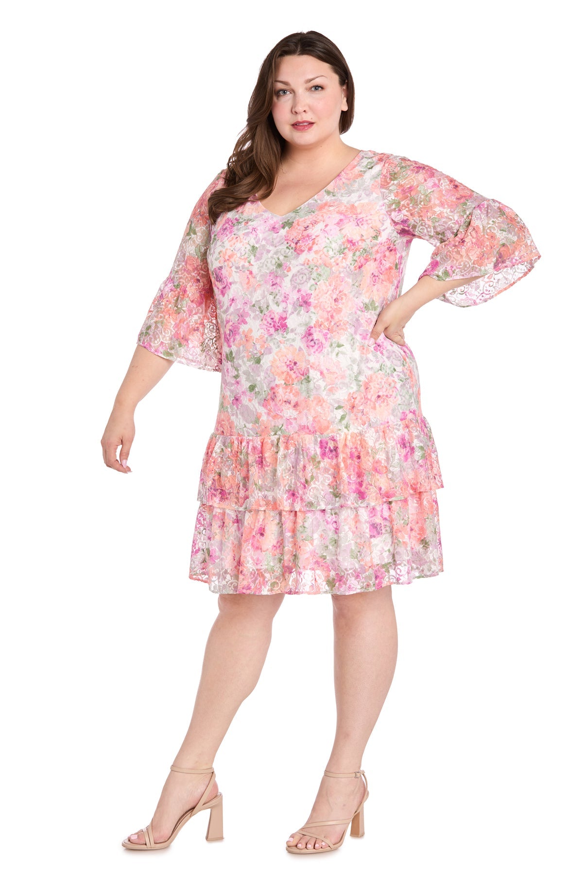 A woman wears the Plus-Size Print Party Dress: R&M Richards 4107W, a pink knee-length floral dress with flared sleeves and a ruffled hem, paired with nude block-heeled sandals, standing against a white background.