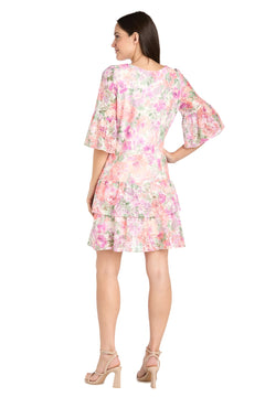 A woman faces away, wearing the Short Ivory Floral Party Dress by R&M Richards (style 4107) featuring ruffled sleeves and a layered skirt, paired with nude high-heeled sandals.